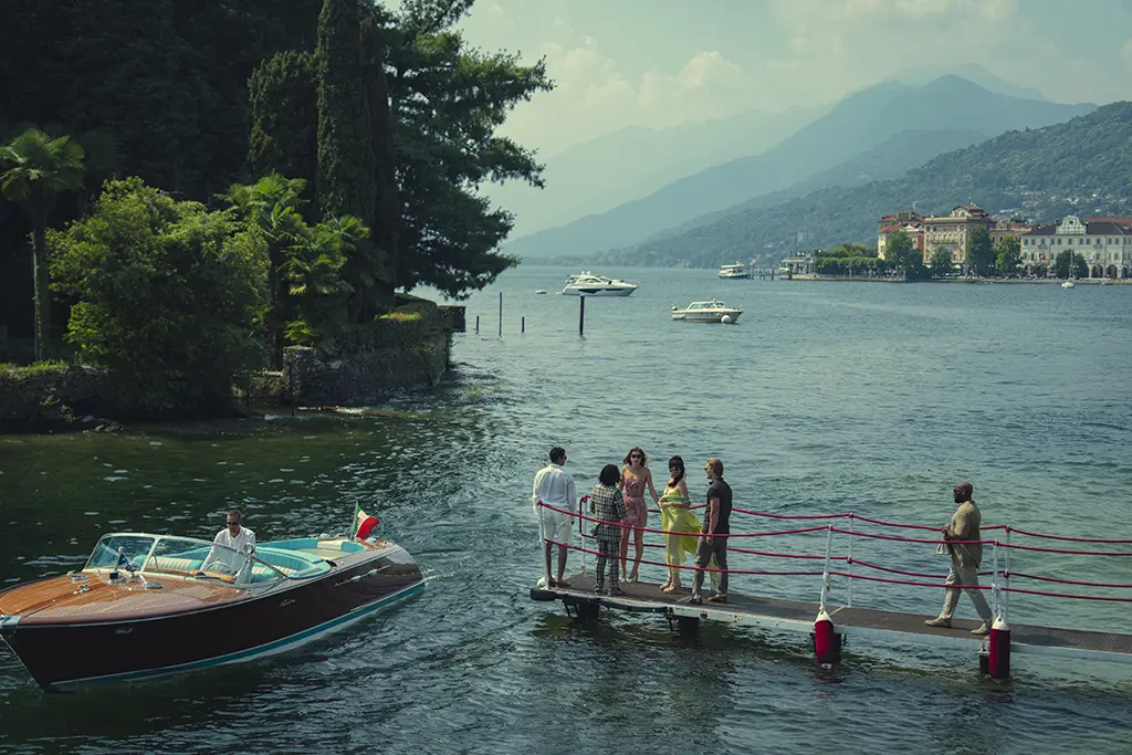 "A group of people stand on a dock by a scenic lake as a wooden motorboat approaches. The background features lush greenery, distant boats, mountains, and elegant lakeside buildings under a hazy sky."