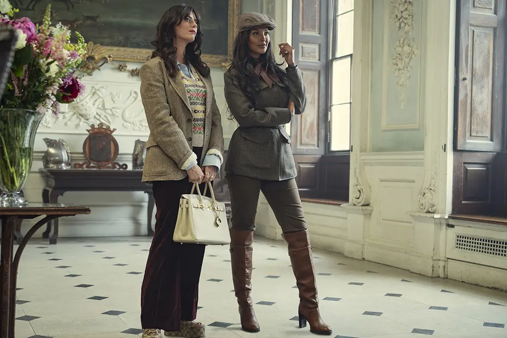 "Two stylish women stand in an elegant, classic interior with tall windows and ornate decor. One holds a white handbag, both wear blazers and boots, appearing poised and fashionable in a well-lit, sophisticated room with flowers and art."